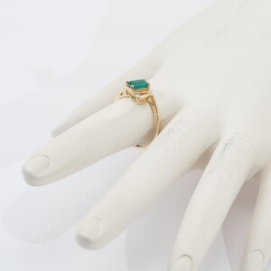May include: A gold ring featuring a rectangular, emerald-cut green gemstone as the primary focal point. The ring has a delicate, ornate design with small accent stones on either side of the emerald. The ring is displayed on a white surface.