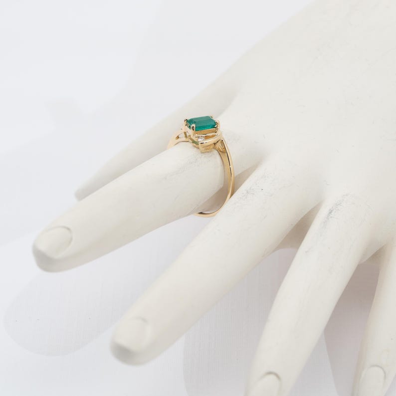 May include: A gold ring featuring a rectangular, emerald-cut green gemstone as the primary focal point. The ring has a delicate, ornate design with small accent stones on either side of the emerald. The ring is displayed on a white surface.