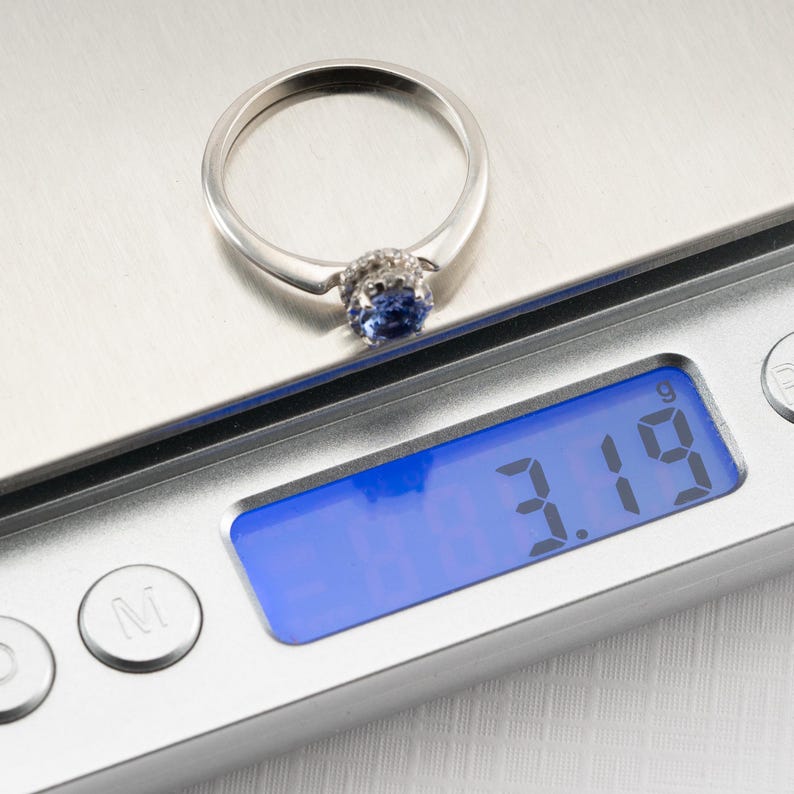 May include: A silver ring with a blue gemstone and small white gemstones around it is placed on a digital scale. The scale reads 3.19 grams.