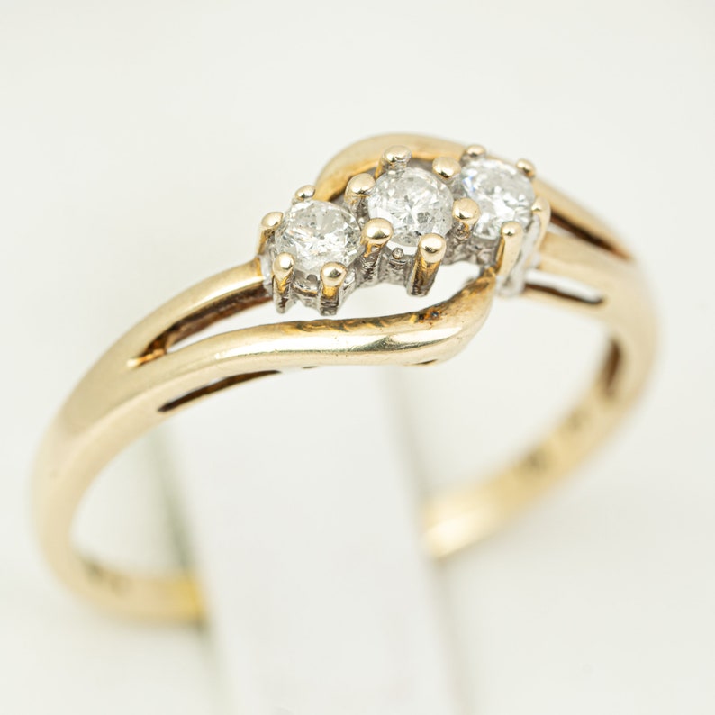 May include: A gold ring featuring three round diamonds set in a row. The band splits into a double-strand design on either side of the diamonds. The ring is set against a white background.
