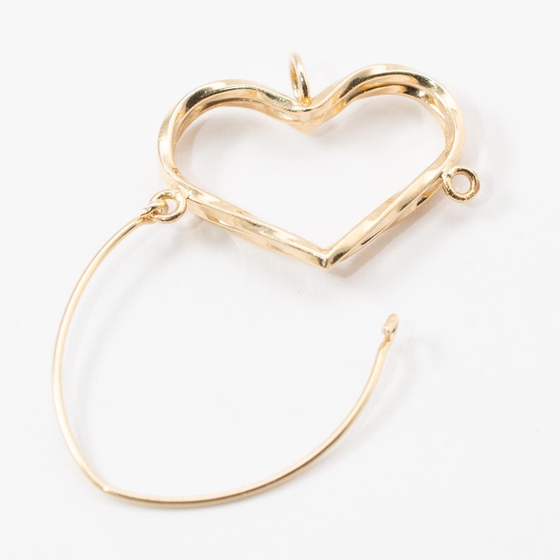Op de afbeelding: Een goudkleurige oorbel met een hartvormige hanger en een gebogen draad. De hartvormige hanger heeft een getextureerd oppervlak en een kleine lus aan de bovenkant. De draad is bevestigd aan de onderkant van het hart en buigt naar buiten.