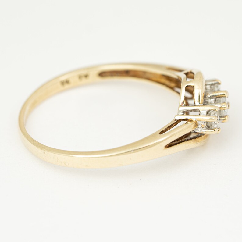 May include: A gold ring with a cluster of small, clear gemstones. The ring has a delicate band and a square-shaped setting for the gemstones. The ring is set against a white background.