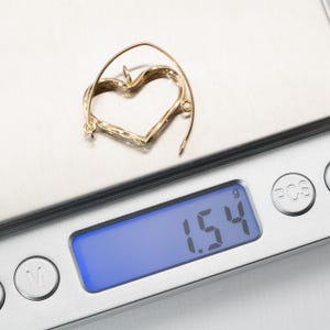 Op de afbeelding: Een goudkleurige oorbel in de vorm van een hart ligt op een digitale weegschaal. De weegschaal geeft een gewicht van 1,54 gram aan. De oorbel heeft een getextureerd oppervlak en een eenvoudig, elegant ontwerp. De achtergrond is een lichtgrijze ondergrond.