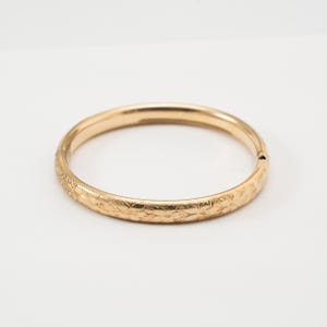 May include: A gold-toned hinged bangle bracelet featuring a continuous engraved floral pattern across its outer surface. The circular piece has a smooth interior and a visible hinge mechanism on the side, maintaining a consistent metallic finish.