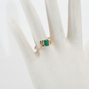 May include: A gold ring featuring a rectangular emerald-cut green gemstone. The ring has a delicate, curved design with small accent stones on either side of the emerald. The ring is displayed on a white surface.
