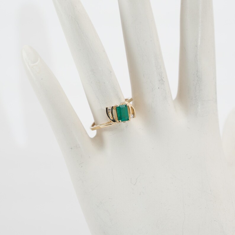 May include: A gold ring featuring a rectangular emerald-cut green gemstone. The ring has a delicate, curved design with small accent stones on either side of the emerald. The ring is displayed on a white surface.