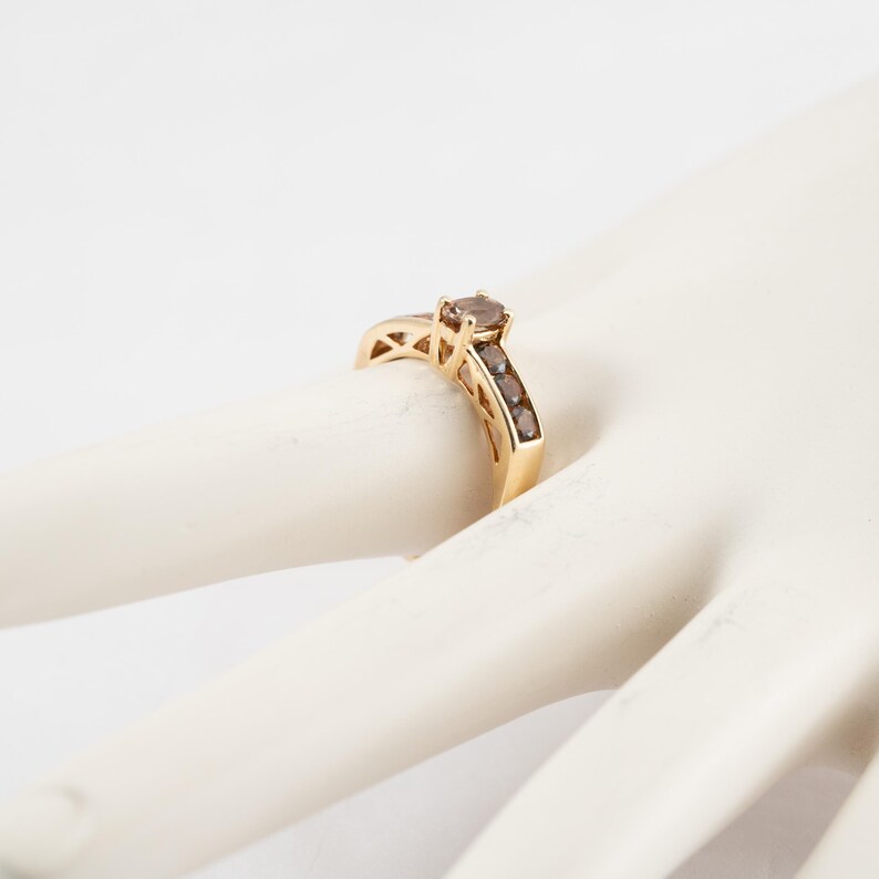 Puede incluir: Un anillo de oro con una piedra preciosa ovalada de color marr&oacute;n en el centro y piedras preciosas marrones m&aacute;s peque&ntilde;as engastadas en una banda alrededor del anillo.