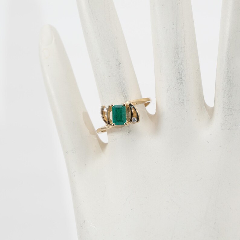 May include: A gold ring featuring a rectangular emerald gemstone as the centerpiece. The ring has a unique design with small accent diamonds on either side of the emerald. The band curves around the finger, creating an elegant and eye-catching piece of jewelry.