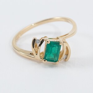 May include: A gold ring featuring a rectangular emerald-cut green gemstone. The ring has a swirling design with small accent diamonds on either side of the emerald. The band is a simple, polished gold.