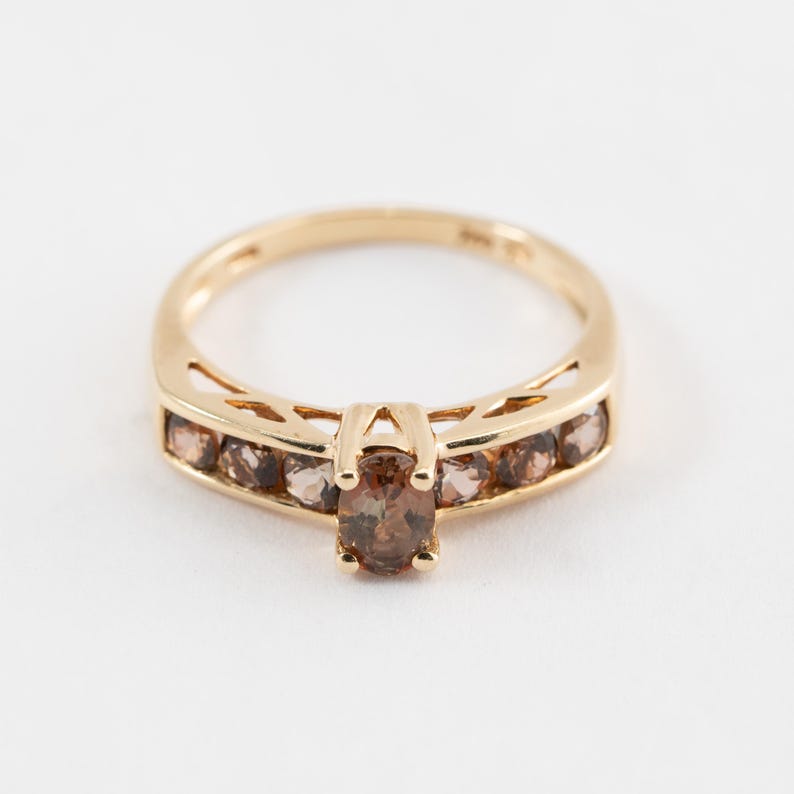 Puede incluir: Un anillo de oro con una piedra preciosa marr&oacute;n en el centro y piedras preciosas marrones m&aacute;s peque&ntilde;as engastadas en una banda alrededor del anillo.