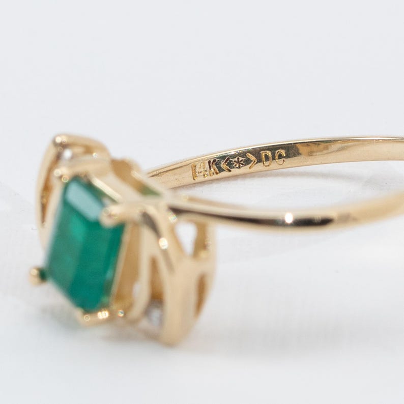May include: Close-up of a gold ring featuring a rectangular, emerald-cut green gemstone. The ring has a delicate, ornate design with small accent stones. The band is stamped with "14K" and other markings.