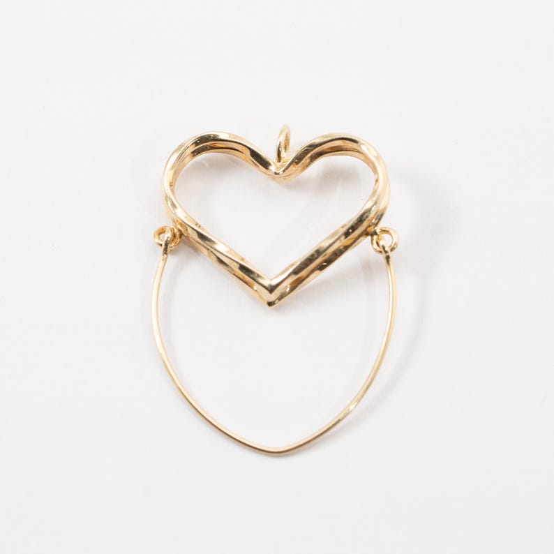Op de afbeelding: Een goudkleurige hartvormige hanger met een getextureerd oppervlak. De hanger heeft een lus aan de bovenkant en een gebogen draad die zich naar beneden uitstrekt, waardoor een uniek ontwerp ontstaat. De sieraden zijn geplaatst tegen een witte achtergrond.