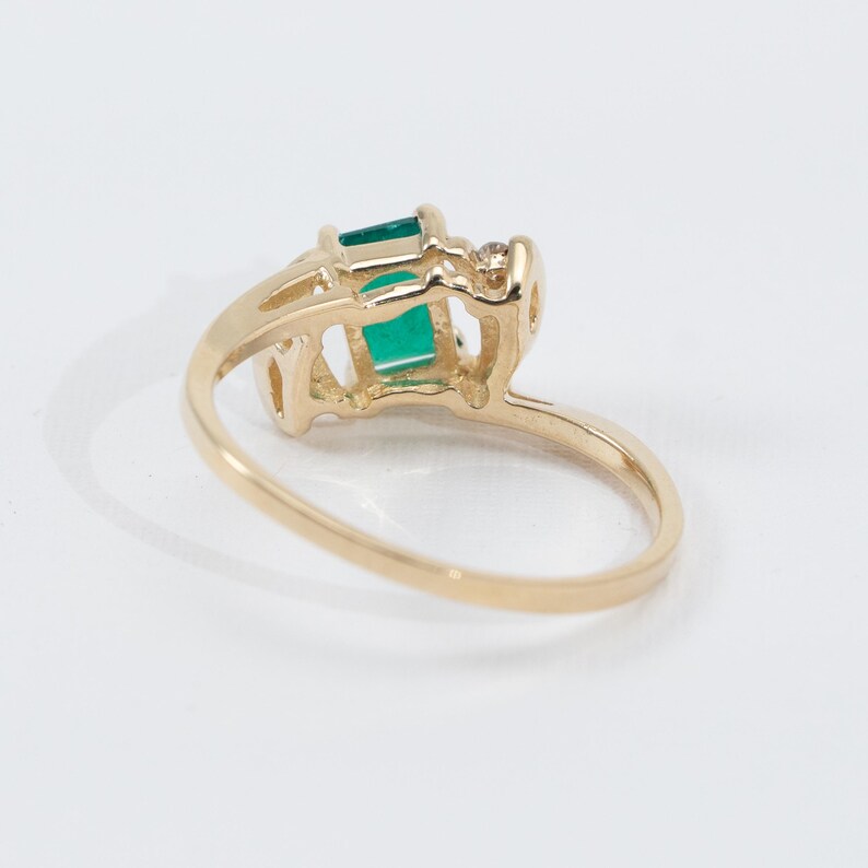 May include: A gold ring featuring a rectangular emerald-cut green gemstone and a small diamond accent. The ring has a delicate, openwork design and a polished finish.