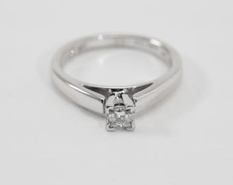 May include: A polished silver-toned metal ring featuring a single, small, square-cut clear gemstone set in a four-prong setting. The band is smooth and tapers slightly toward the center, where the stone is securely held in a raised, open-sided mount.
