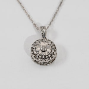 May include: A silver pendant necklace with a round, diamond-studded pendant. The pendant has a central starburst design surrounded by multiple rows of smaller diamonds. The chain is a delicate silver cable chain.