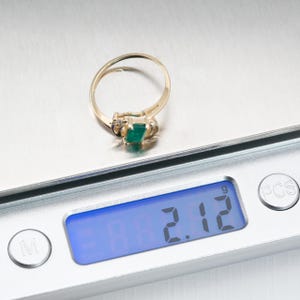 May include: A gold ring with a rectangular green gemstone is displayed on a digital scale. The scale shows a weight of 2.12. The ring has a delicate band and is likely a piece of jewelry.