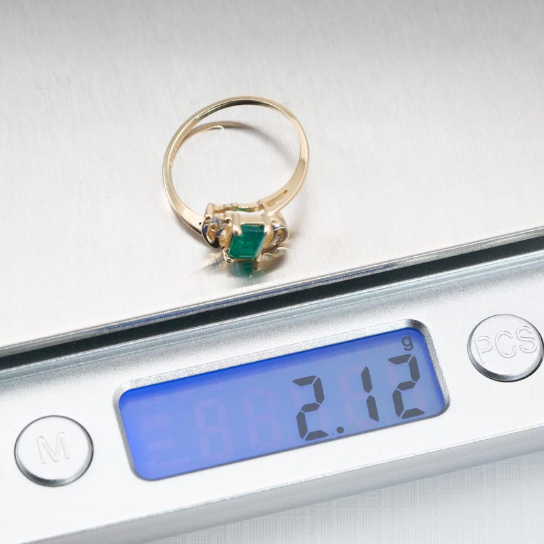 May include: A gold ring with a rectangular green gemstone is displayed on a digital scale. The scale shows a weight of 2.12. The ring has a delicate band and is likely a piece of jewelry.
