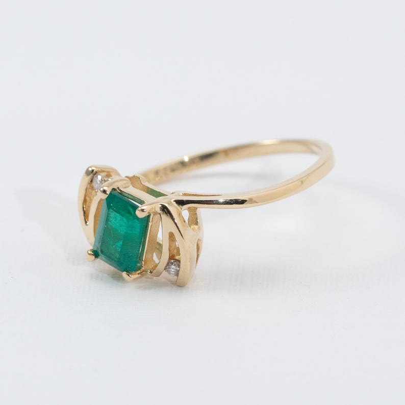 May include: A gold ring featuring a square-cut emerald gemstone as the primary focal point. The ring has a delicate, openwork design on the sides and small accent diamonds. The band is a classic, polished gold.