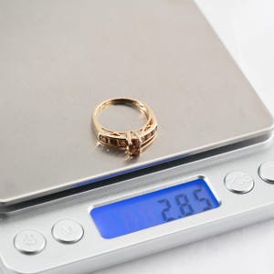Puede incluir: Un anillo de oro con piedras preciosas marrones en una b&aacute;scula digital que marca 2,85 gramos.