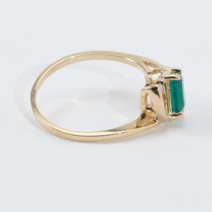 May include: A gold ring featuring a rectangular, emerald-cut green gemstone. The ring has a delicate, openwork design on the sides of the gemstone setting. The band is a simple, polished gold.