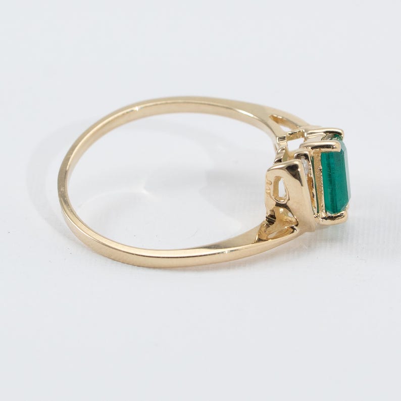 May include: A gold ring featuring a rectangular, emerald-cut green gemstone. The ring has a delicate, openwork design on the sides of the gemstone setting. The band is a simple, polished gold.