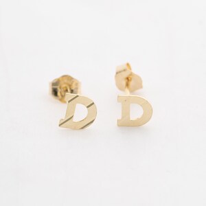 May include: A pair of gold initial earrings featuring the letter "D". The earrings are made of solid gold and have a polished finish.