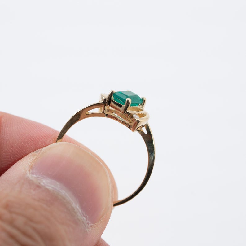 May include: A gold ring featuring a rectangular, emerald-cut green gemstone. The ring has a delicate, curved band and ornate detailing around the gemstone setting. The ring is held against a white background.