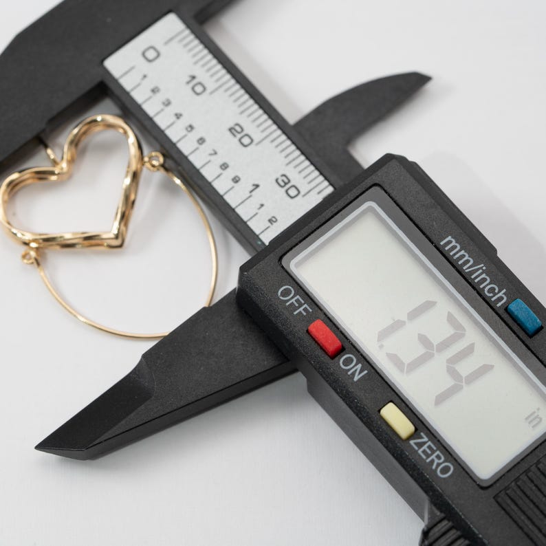 Op de afbeelding: Een goudkleurige hartvormige oorring wordt gemeten met een digitale schuifmaat. De schuifmaat geeft een meting van 3,4 cm weer. De oorring heeft een getextureerd hartontwerp en een gladde, cirkelvormige ring.