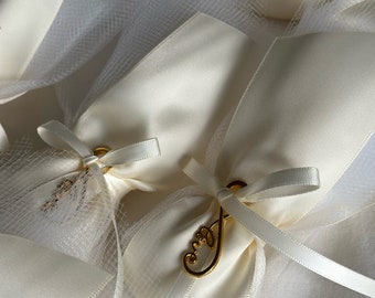 May include: White satin favor bags with gold ribbon and a gold charm that says "Love" on it. The bags are tied with a bow and are sitting on a white tulle fabric.