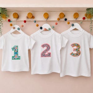 May include: Three white short-sleeved t-shirts hanging on white hangers. The first shirt features a number one with a blue dinosaur print, the second a number two with a pink floral pattern, and the third a number three with a colourful floral print.