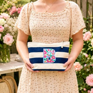 May include: A rectangular canvas pouch featuring horizontal navy blue and cream stripes. The center of the pouch displays a large, embroidered letter B filled with a vibrant floral pattern in shades of pink, green, and blue, finished with a zipper.