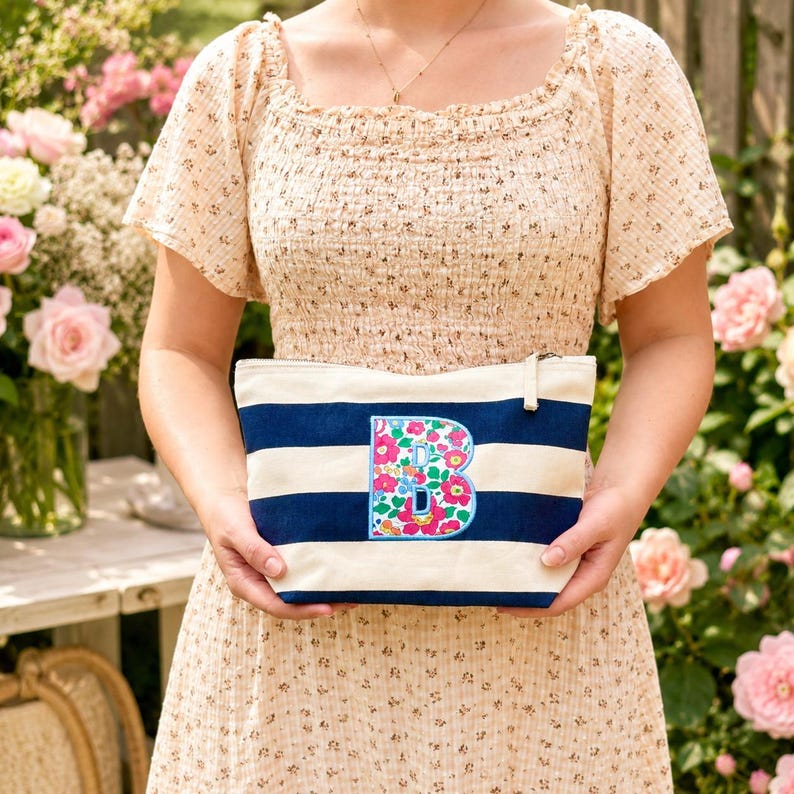May include: A rectangular canvas pouch featuring horizontal navy blue and cream stripes. The center of the pouch displays a large, embroidered letter B filled with a vibrant floral pattern in shades of pink, green, and blue, finished with a zipper.
