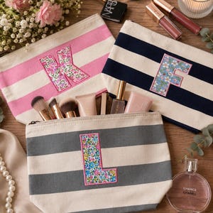 May include: Three canvas zipper pouches featuring horizontal stripes in pink, navy blue, and gray. Each pouch displays a large, centered letter&mdash;K, F, or L&mdash;appliqu&eacute;d in a colorful floral fabric with a pink stitched border. The pouches are arranged on a wooden surface alongside various makeup brushes, cosmetics, a candle, and a string of pearls.