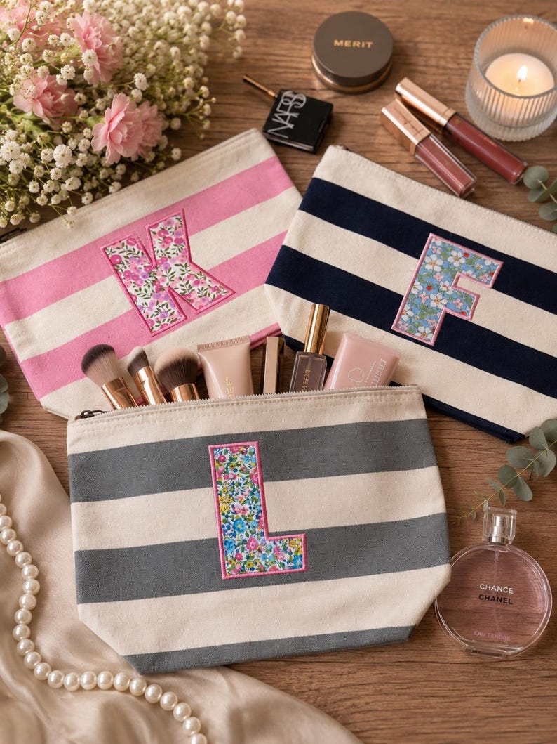 May include: Three canvas zipper pouches featuring horizontal stripes in pink, navy blue, and gray. Each pouch displays a large, centered letter&mdash;K, F, or L&mdash;appliqu&eacute;d in a colorful floral fabric with a pink stitched border. The pouches are arranged on a wooden surface alongside various makeup brushes, cosmetics, a candle, and a string of pearls.