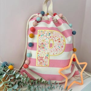 May include: A pink and white striped drawstring bag featuring a large embroidered letter "P" with a floral and star design. The bag has a cream-coloured drawstring and handle. A colourful pom-pom garland and an orange star decoration are also present.