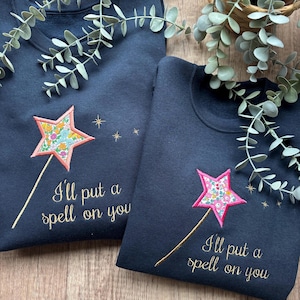 May include: Two navy blue crewneck sweatshirts featuring embroidered designs. Each sweatshirt displays a star-shaped wand with a colourful floral pattern and gold stitching. The text "I'll put a spell on you" is embroidered in gold cursive script.