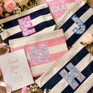 May include: A collection of rectangular canvas zipper pouches featuring horizontal stripes in navy blue and white or pink and white. Each pouch displays a large, fabric-appliqued letter with a floral or dinosaur pattern. A card reads Thank You for being an amazing teacher.