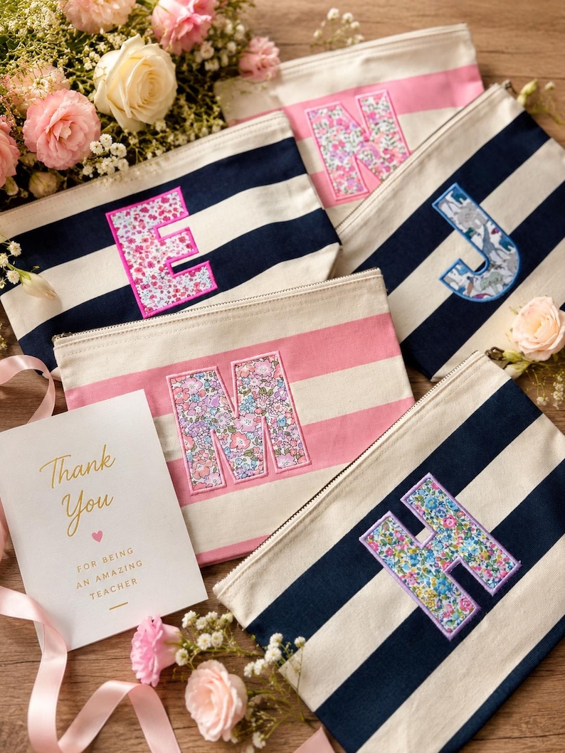 May include: A collection of rectangular canvas zipper pouches featuring horizontal stripes in navy blue and white or pink and white. Each pouch displays a large, fabric-appliqued letter with a floral or dinosaur pattern. A card reads Thank You for being an amazing teacher.
