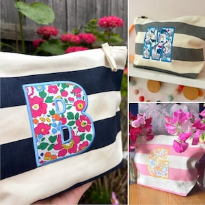 May include: A collection of three zippered pouches featuring horizontal stripes and large fabric letters appliqued on the front. One pouch has navy stripes with a floral letter B, one has grey stripes with an animal print letter M, and one has pink stripes with a floral letter G.