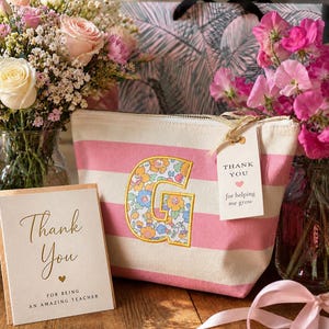 May include: A canvas pouch with pink and white horizontal stripes featuring a large letter G appliqued in a floral pattern. A white tag attached to the gold zipper reads Thank You for helping me grow, alongside a card reading Thank You for being an amazing teacher.