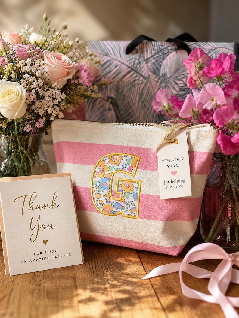 May include: A canvas pouch with pink and white horizontal stripes featuring a large letter G appliqued in a floral pattern. A white tag attached to the gold zipper reads Thank You for helping me grow, alongside a card reading Thank You for being an amazing teacher.