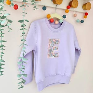 K&ouml;nnte beinhalten: Ein lavendelfarbener Pullover mit einem floralen Muster in Form des Buchstabens E auf der Vorderseite. Der Pullover h&auml;ngt an einem Kleiderb&uuml;gel mit bunten Pompons im Hintergrund.