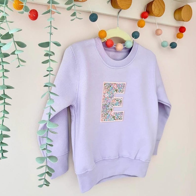 K&ouml;nnte beinhalten: Ein lavendelfarbener Pullover mit einem floralen Muster in Form des Buchstabens E auf der Vorderseite. Der Pullover h&auml;ngt an einem Kleiderb&uuml;gel mit bunten Pompons im Hintergrund.