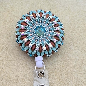May include: A teal and brown beaded badge holder with a clear plastic retractable reel. The beaded portion is round and features a crystal in the center.