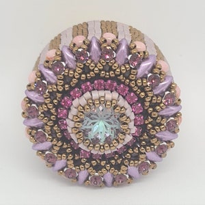 May include: A beaded ring with a pink, white and gold colour scheme. The ring features a large, iridescent crystal in the centre surrounded by smaller pink crystals.