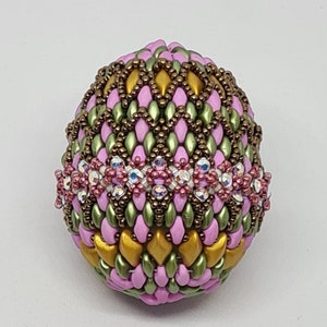 May include: A decorative beaded egg in shades of pink, green, and gold. The egg is intricately designed with a pattern of small beads and a band of crystals. The egg is likely a handcrafted item, suitable for display or as a unique gift.