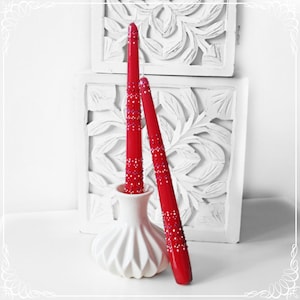 Könnte beinhalten: Zwei rote Kerzen mit einem glitzernden Design werden in einer weißen Keramikvase präsentiert. Die Kerzen sind hoch und schlank, und die Vase hat ein strukturiertes, plissiertes Design.