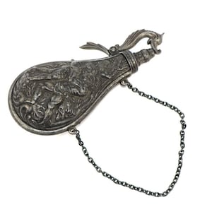Pode incluir: Um frasco de pólvora de metal prateado com um relevo intrincado de uma figura humana no corpo. O frasco tem um gargalo estreito com um mecanismo de bico articulado e uma fina corrente de metal presa na lateral para transporte ou fixação.