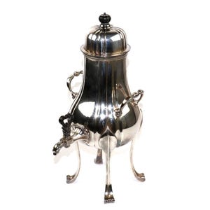 Pode incluir: Uma cafeteira antiga prateada com tampa abobadada e um botão preto. Possui duas alças, uma torneira e assenta em três pernas curvas. A superfície polida reflete a luz, destacando o seu design ornamentado. Uma peça clássica.