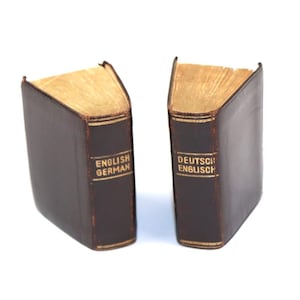 May include: Two vintage bookends in a dark brown leather-like material. The bookends have gold-colored gilded edges and spines. One bookend reads "ENGLISH GERMAN" and the other reads "DEUTSCH ENGLISCH" in gold lettering. The bookends are on a white background.